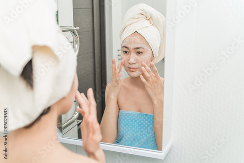 Asian girl in a mask with a towel in front of the mirror in the bathroom. the concept of self-care, moisturizing and cleansing the skin. care of youth and beauty