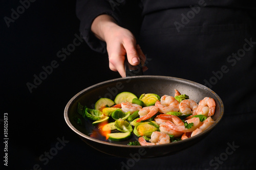 Chef cooks with fire in a pan shrimp with vegetables. On a black background, restaurant service