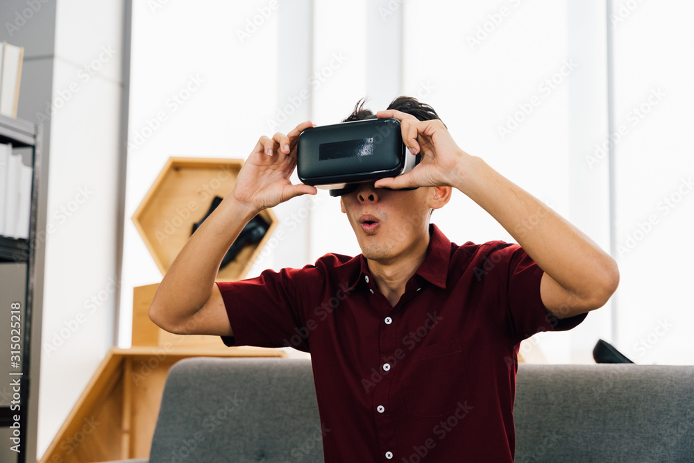 Amazed young Asian man in casual clothes with open mouth playing video games wearing VR goggles sitting on sofa at home Amazed young Asian man in casual clothes with open mouth playing video games wearing VR goggles sitting on sofa at home
