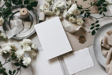 Moody wedding table mockup scene. Feminine desktop composition with fading white rose flowers, silver plate, silk ribbons, seal stamp, envelopes and blank greeting cards. Grey background. flat lay,top