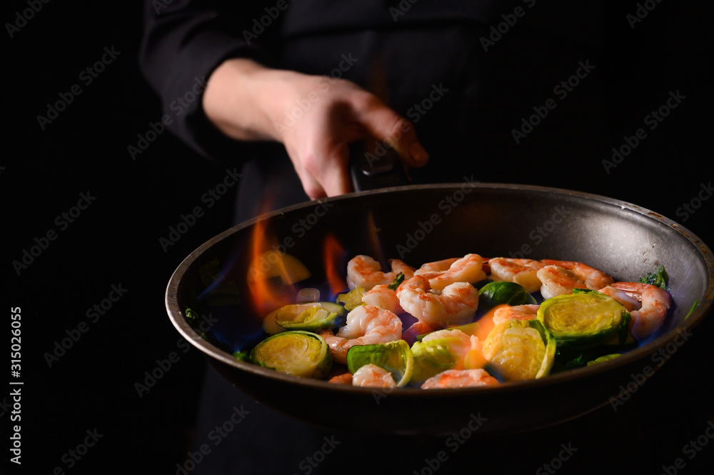 Chef cooks with fire in a pan shrimp with vegetables. On a black background, restaurant service Chef cooks with fire in a pan shrimp with vegetables. On a black background, restaurant service