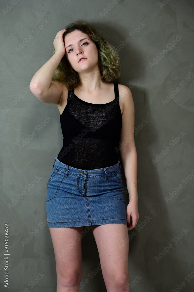 Concept vertical photo of a young woman in a black T-shirt and blue skirt standing straight. Portrait of a pretty student girl with beautiful curly hair in front of the camera on a gray background. Concept vertical photo of a young woman in a black T-shirt and blue skirt standing straight. Portrait of a pretty student girl with beautiful curly hair in front of the camera on a gray background.
