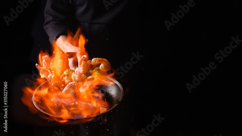 Seafood and their preparation, a professional chef cooks shrimps with vegetables with fire. On a black background, a banner, cooking and recipes. Restaurant and hotel service concept.