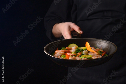 Professional chef cook on a dark background prepares seafood, shrimp with fire and vegetables. Roasting and cooking veggie food. Black background, Space for design.