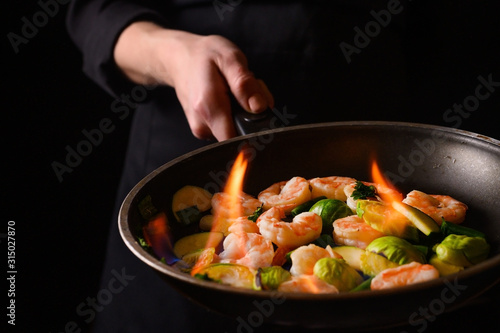 Chef cooks with fire in a pan shrimp with vegetables. On a black background, restaurant service