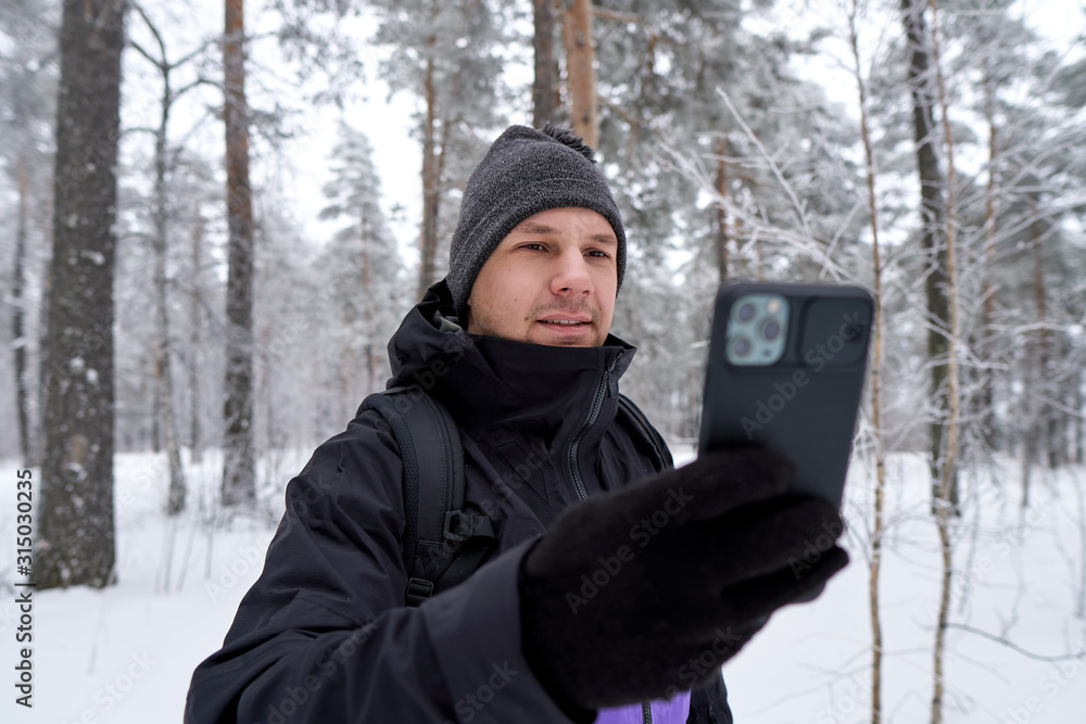 Young man hiker takes photo selfie on new smartphone; high steep snowy stone slopes of mountains and pine fir forest in snowy winter day on background; tourist look at the phone; mobile navigation Young man hiker takes photo selfie on new smartphone; high steep snowy stone slopes of mountains and pine fir forest in snowy winter day on background; tourist look at the phone; mobile navigation