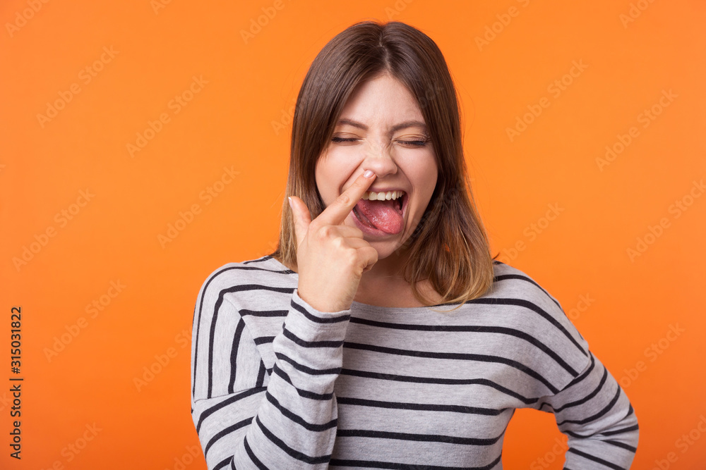 Φωτογραφία Portrait of funny joyful young woman with brown hair in long sleeve striped shirt standing, picking nose and sticking her tongue out, foolish grimace