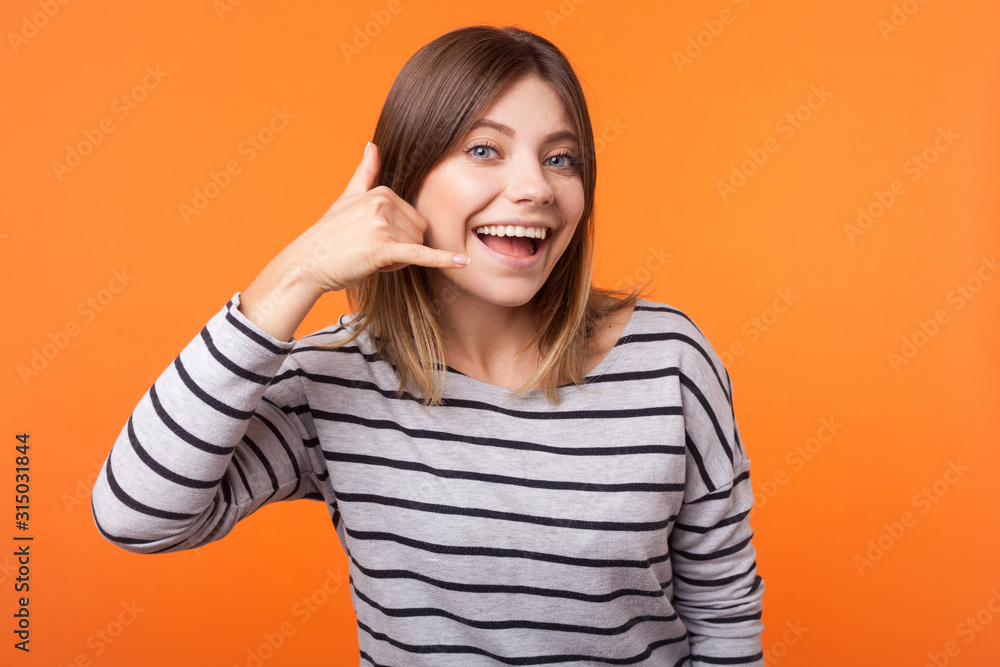 Call me! Portrait of cheerful beautiful woman with brown hair in long sleeve striped shirt standing, making call gesture with hand, looking playful. indoor studio shot isolated on orange background Call me! Portrait of cheerful beautiful woman with brown hair in long sleeve striped shirt standing, making call gesture with hand, looking playful. indoor studio shot isolated on orange background