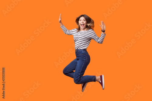 Full length portrait of extremely happy pretty woman with brown hair in long sleeve shirt and denim jumping for joy or flying, celebrating success. indoor studio shot isolated on orange background