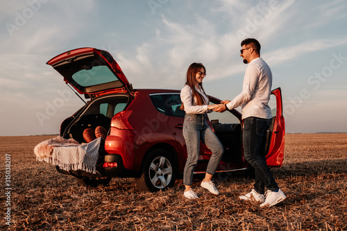 Young Happy Couple Dressed Alike in White Shirt and Jeans Enjoying Road Trip at Their New Car, Beautiful Sunset on the Field, Vacation and Travel Concept