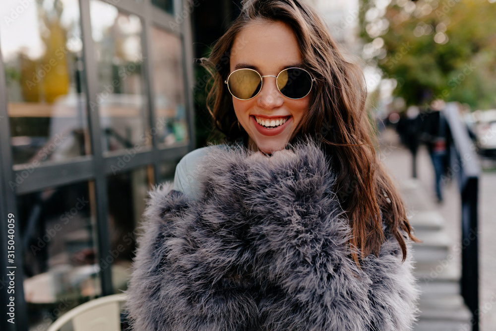 Close up outdoor portrait of laughing woman with wavy hair wearing sunglasses and fur coat looking at camera with wonderful smile Close up outdoor portrait of laughing woman with wavy hair wearing sunglasses and fur coat looking at camera with wonderful smile
