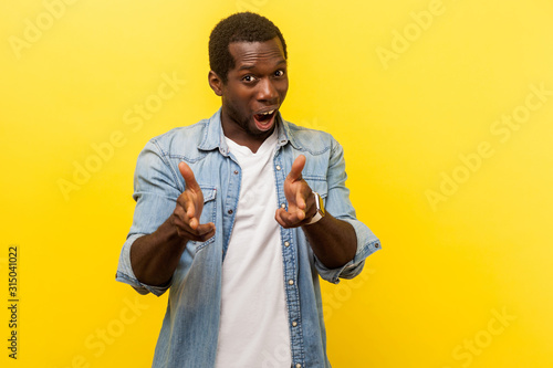 Portrait of street criminal, dangerous young man in denim casual shirt pointing with finger pistols, standing with open mouth and threatening to shoot. indoor studio shot isolated on yellow background