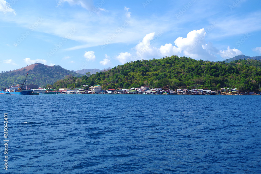 A small, colorful town hugging the coast of an island in the Philippines archipelago. It's a beautiful, sunny, summer day and the waters of the ocean are very blue and calm.  