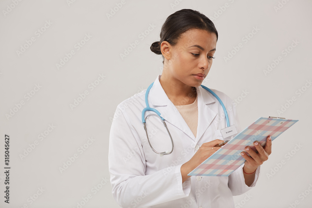 Waist up portrait of young mixed race nurse or female doctor holding clipboard while standing against white background, copy space Waist up portrait of young mixed race nurse or female doctor holding clipboard while standing against white background, copy space