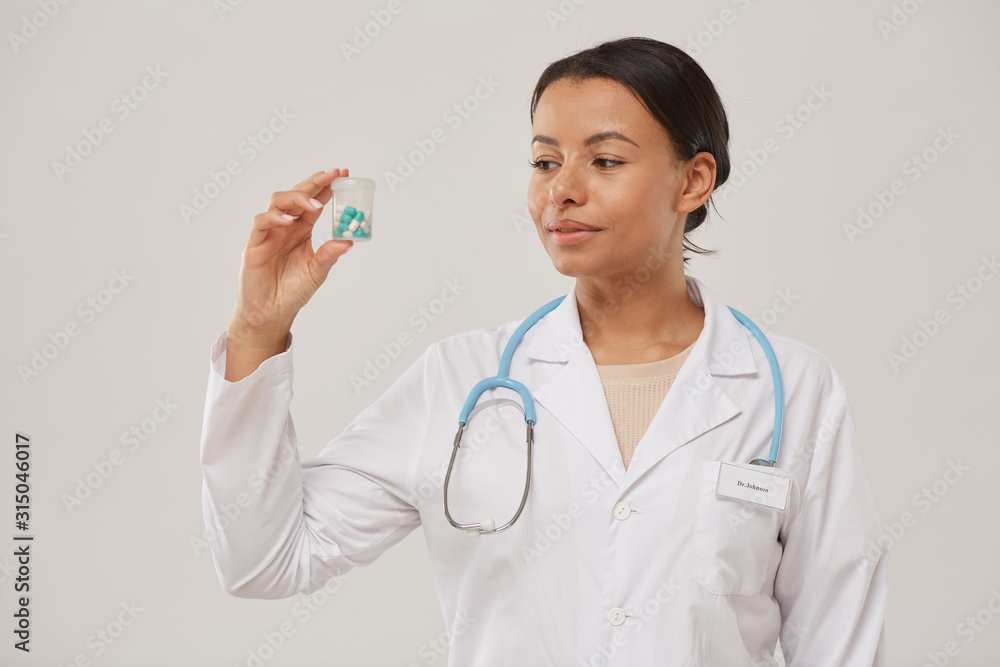 Waist up portrait of beautiful female doctor holding pill bottle while standing against white background, copy space Waist up portrait of beautiful female doctor holding pill bottle while standing against white background, copy space