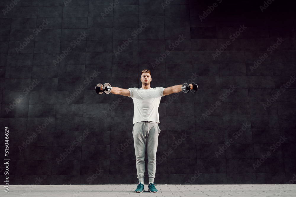 Full length of attractive caucasian muscular bearded blonde man in tracksuit and with t-shirt standing in front of gray wall and holding dumbbells.