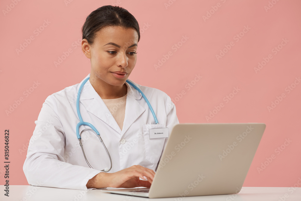 Portrait of beautiful female doctor using laptop while sitting against pale pink background, copy space Portrait of beautiful female doctor using laptop while sitting against pale pink background, copy space