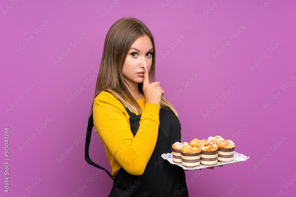 Teenager girl holding lots of different mini cakes over isolated purple background doing silence gesture Teenager girl holding lots of different mini cakes over isolated purple background doing silence gesture