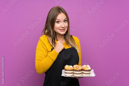 Teenager girl holding lots of different mini cakes over isolated purple background celebrating a victory