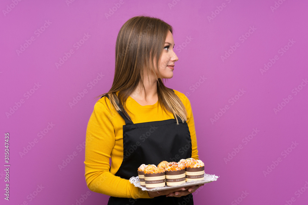 Teenager girl holding lots of different mini cakes over isolated purple background looking to the side Teenager girl holding lots of different mini cakes over isolated purple background looking to the side