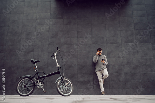 Full length of handsome Caucasian fashionable man leaning on gray wall and using smart phone for reading or sending message. In foreground is bicycle. © dusanpetkovic1