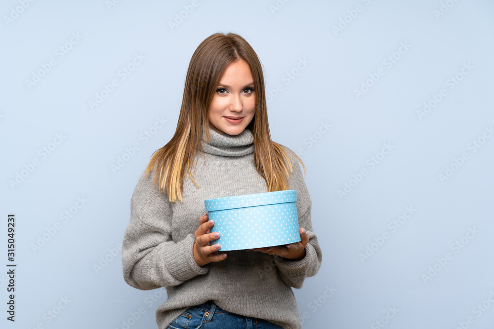 Teenager girl with sweater over isolated blue background holding gift box Teenager girl with sweater over isolated blue background holding gift box