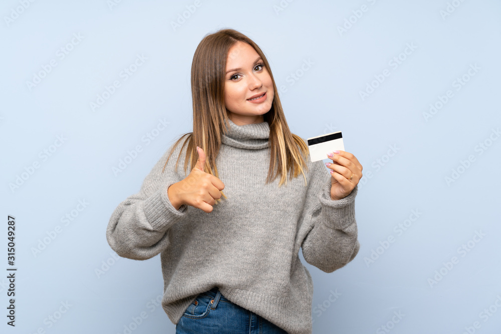 Teenager girl with sweater over isolated blue background holding a credit card Teenager girl with sweater over isolated blue background holding a credit card