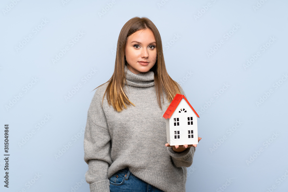 Teenager girl with sweater over isolated blue background holding a little house Teenager girl with sweater over isolated blue background holding a little house