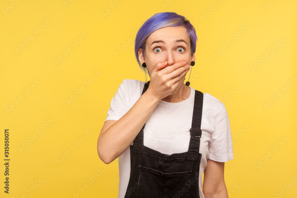 Portrait of scared terrified hipster girl with violet short hair in denim overalls covering mouth with hand and looking intimidated with eyes full of fear. isolated on yellow background, studio shot Portrait of scared terrified hipster girl with violet short hair in denim overalls covering mouth with hand and looking intimidated with eyes full of fear. isolated on yellow background, studio shot