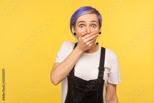 Portrait of scared terrified hipster girl with violet short hair in denim overalls covering mouth with hand and looking intimidated with eyes full of fear. isolated on yellow background, studio shot