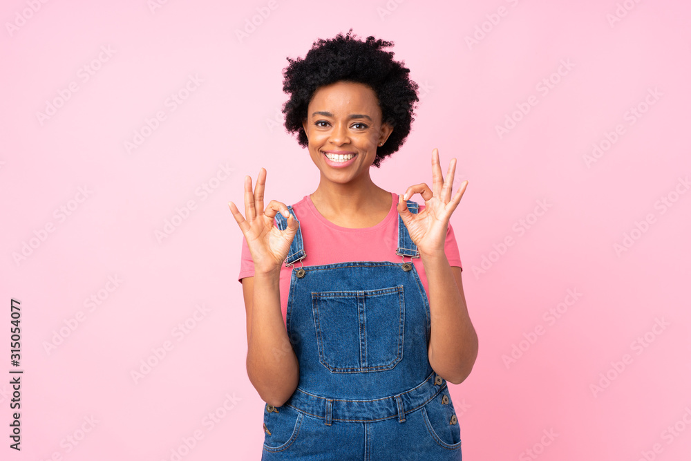 African american woman with overalls over isolated pink background showing an ok sign with fingers African american woman with overalls over isolated pink background showing an ok sign with fingers