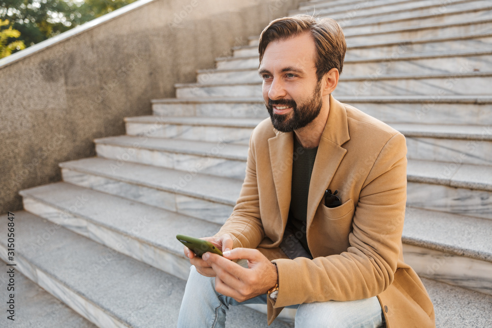 Obraz premium Image of handsome man holding smartphone while sitting on stairs outdoors