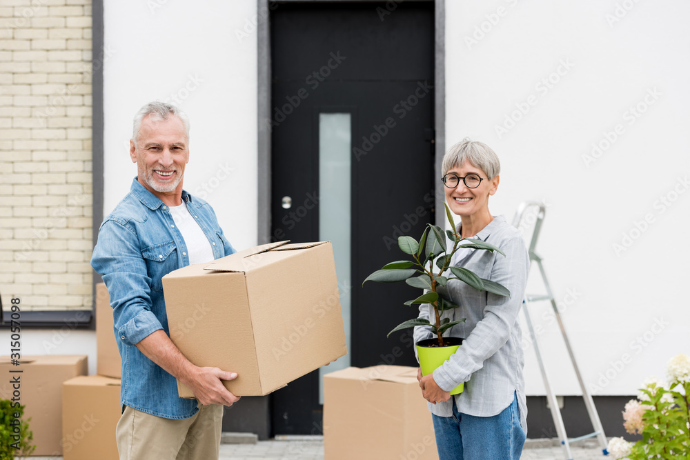 mature man holding box and smiling woman holding plant near new house mature man holding box and smiling woman holding plant near new house