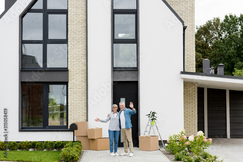 smiling mature man and woman waving near new house