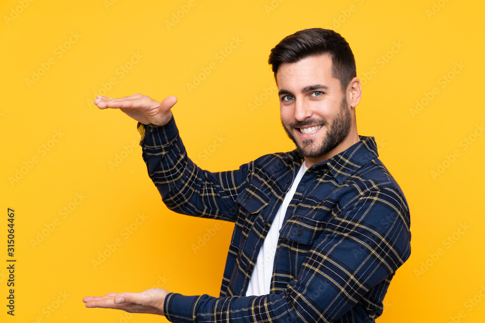 Young handsome man with beard over isolated yellow background holding copyspace to insert an ad Young handsome man with beard over isolated yellow background holding copyspace to insert an ad