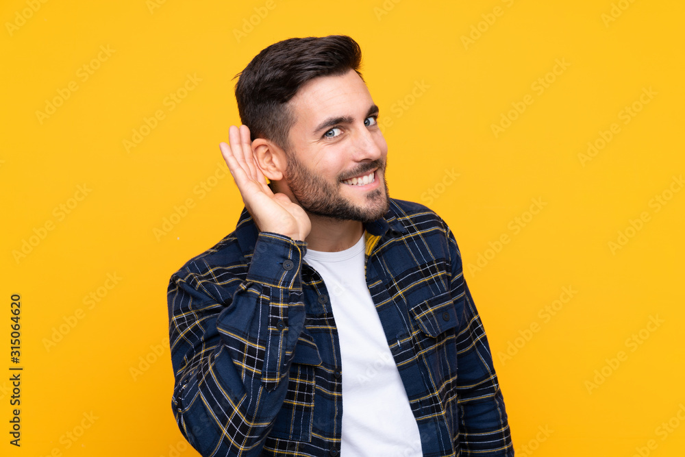 Young handsome man with beard over isolated yellow background listening to something by putting hand on the ear Young handsome man with beard over isolated yellow background listening to something by putting hand on the ear