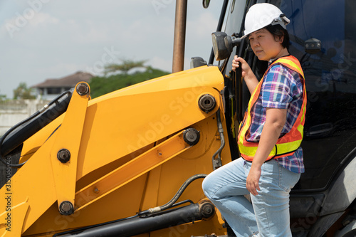 Woman Engineering wearing a white safety helmet standing In front of the backhoe And are using tablet to check the blueprint with construction