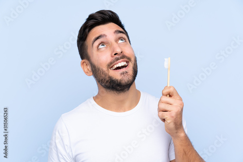 Young handsome man with beard brushing his teeth over isolated background looking up while smiling