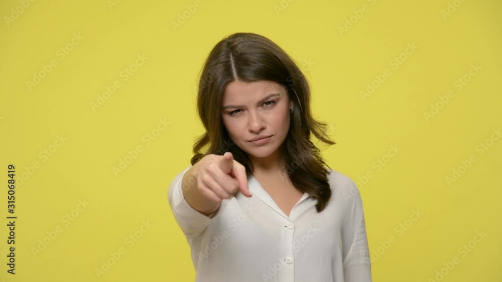 Hey you! Beautiful young displeased woman in blouse looking suspiciously and pointing finger at camera, choosing you and looking serious and strict. indoor studio shot isolated on yellow background Hey you! Beautiful young displeased woman in blouse looking suspiciously and pointing finger at camera, choosing you and looking serious and strict. indoor studio shot isolated on yellow background