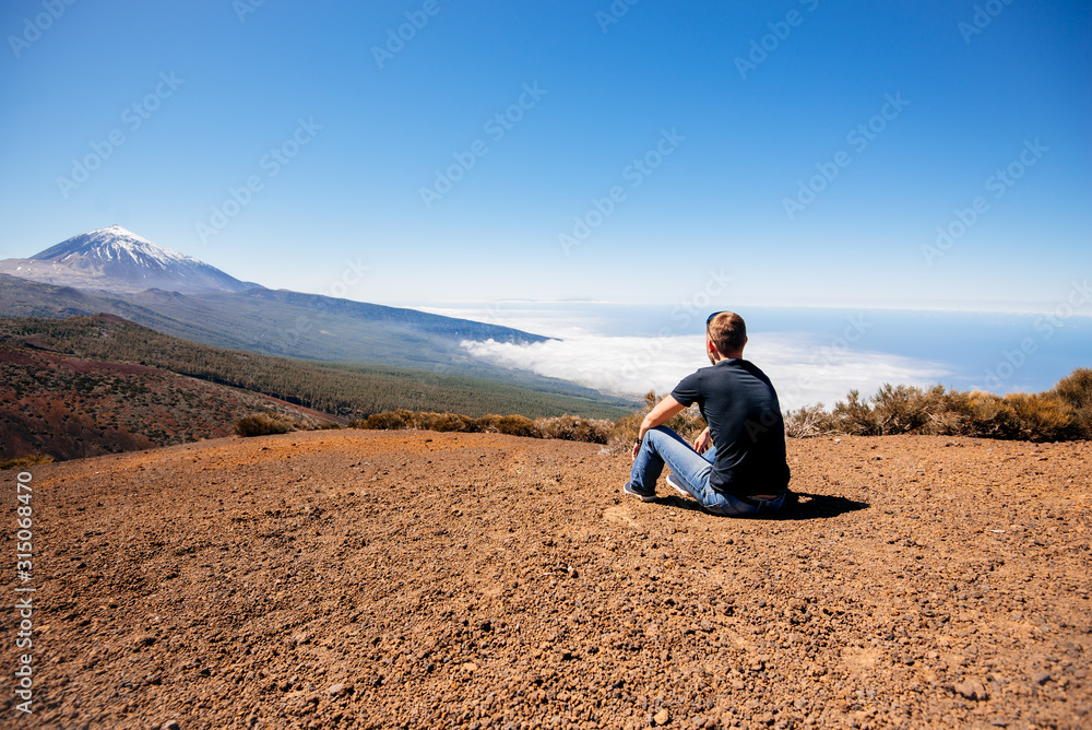 A young male traveler sits on the sand and looks at the top of the Teide volcano. Teide National Park, Canary, Spain