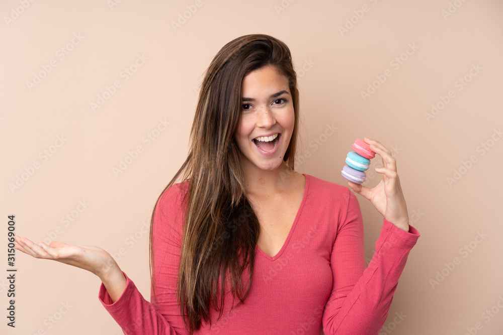 Teenager girl over isolated background holding colorful French macarons with shocked expression Teenager girl over isolated background holding colorful French macarons with shocked expression