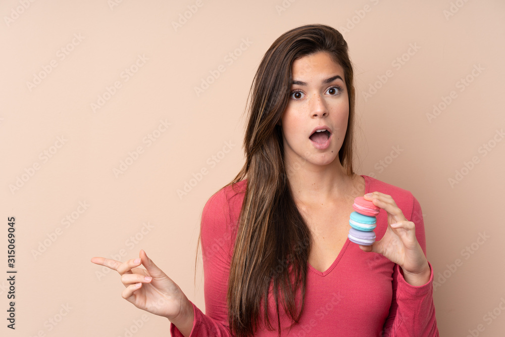 Teenager girl over isolated background holding colorful French macarons and pointing side Teenager girl over isolated background holding colorful French macarons and pointing side