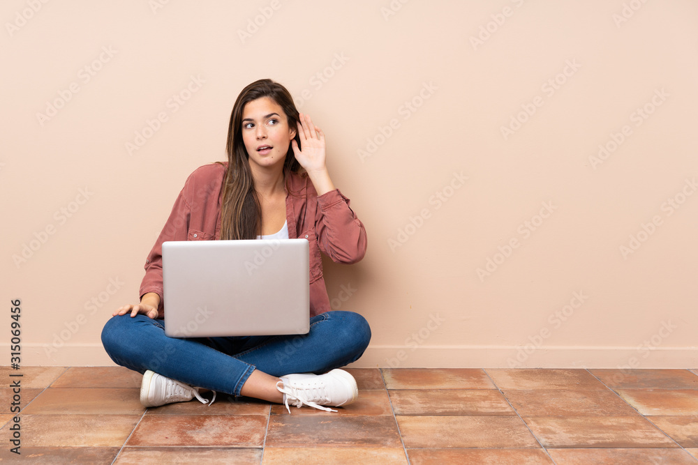 Teenager student girl sitting on the floor with a laptop listening to something by putting hand on the ear Teenager student girl sitting on the floor with a laptop listening to something by putting hand on the ear