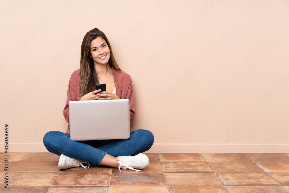 Teenager student girl sitting on the floor with a laptop sending a message with the mobile Teenager student girl sitting on the floor with a laptop sending a message with the mobile