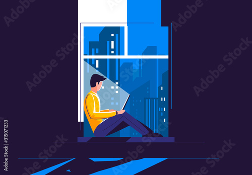 A young man is sitting on the windowsill with a laptop, in the dark against the background of the night city outside the window