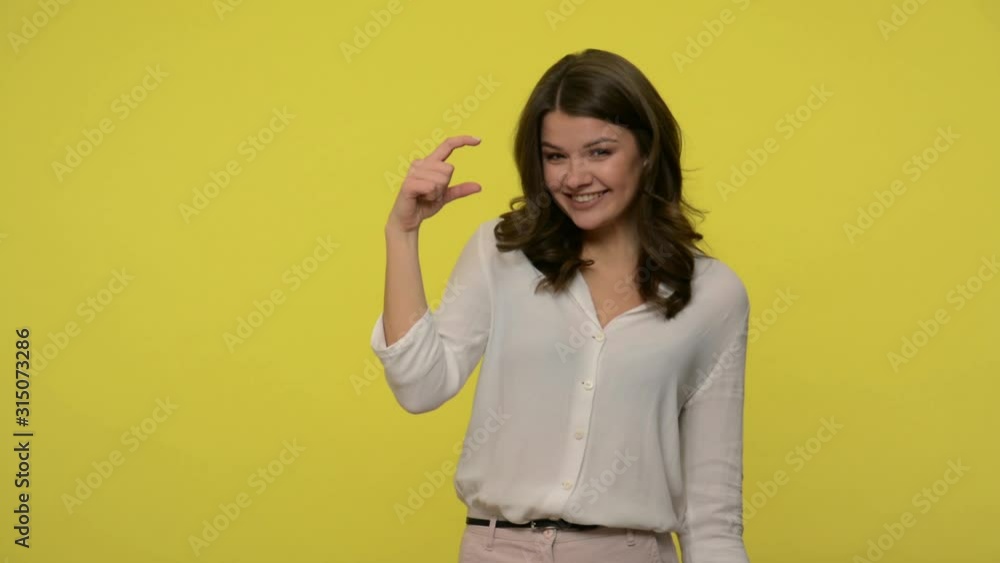 Positive attractive woman with brunette hair in blouse showing little bit sign and smiling playfully at camera, making small size gesture with fingers. indoor studio shot isolated on yellow background Positive attractive woman with brunette hair in blouse showing little bit sign and smiling playfully at camera, making small size gesture with fingers. indoor studio shot isolated on yellow background