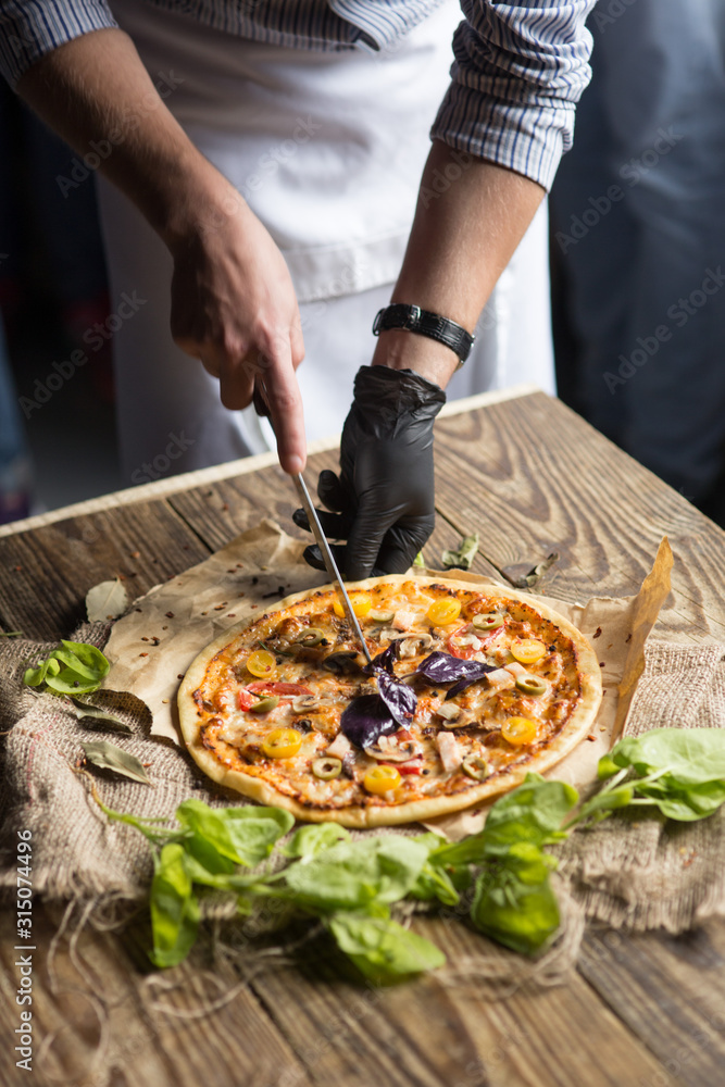 Pizzaiolo cutting with knife pizza with olives basilic mozarella mushrooms on a wooden table and green basil around
