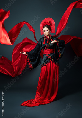 Woman is dressed in red Chinese Japanese folk clothing. Flying fabric, beautiful umbrella and fan in Japanese Chinese style, long earrings in the ears. Girl posing on a dark background