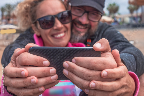 Two couple of hands of a middle-aged couple holding the mobile phone and smile for a selfie. Two attractive and relaxed people. Sitting on the beach on a winter vacation © luciano