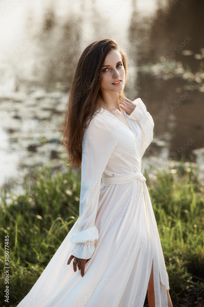 Beautiful slender woman in a long white dress walks in the morning near the lake. Brown-haired girl with long hair walks on the grass in the village, natural cosmetics and makeup Beautiful slender woman in a long white dress walks in the morning near the lake. Brown-haired girl with long hair walks on the grass in the village, natural cosmetics and makeup